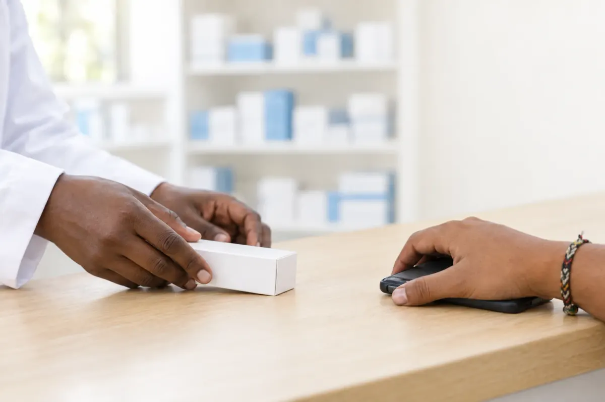 A pharmacist slides a medicine box across the counter to a patient holding a phone